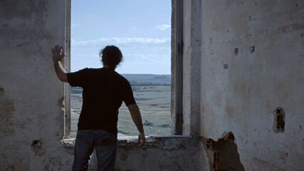 The ruins of an old abandoned house, with a man near a window, looking outside. Establishing shot.
