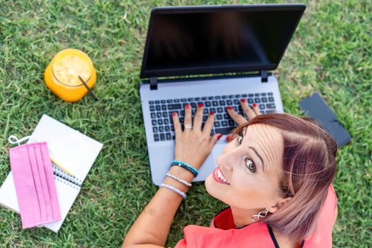 Woman 40 Years Old Working Outdoors With Laptop While Making Healthy Breakfast - Smiling Female Entrepreneur Or Influencer Lying Down On Lawn Typing On Pc During End Of Coronavirus Outbreak - Top View