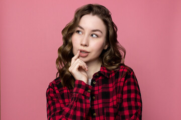 Pensive young woman isolated on pink background. Thoughtful caucasian girl in red shirt with curly hairstyle. Confused woman