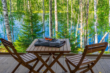 View from panoramic cottage window to outdoor terrace with table and chairs against beautiful landscape of lake in northern forest. Glasses with alcohol drink wrapped in sweater on table. Staycation