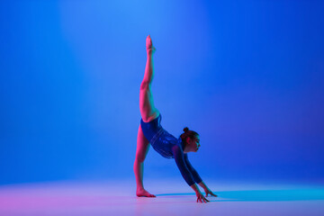 Agility. Young flexible girl isolated on blue studio background in neon light. Young female model practicing artistic gymnastics. Exercises for flexibility, balance. Grace in motion, sport, action.