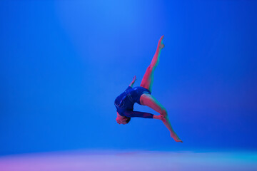 Flying high. Young flexible girl isolated on blue studio background in neon light. Young female model practicing artistic gymnastics. Exercises for flexibility, balance. Grace in motion, sport, action