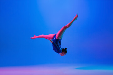 Fototapeta na wymiar Flying high. Young flexible girl isolated on blue studio background in neon light. Young female model practicing artistic gymnastics. Exercises for flexibility, balance. Grace in motion, sport, action