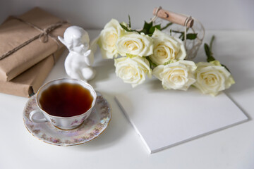A porcelain tea cup with a saucer, a teapot, a white faience angel and three roses on the table. Breakfast.