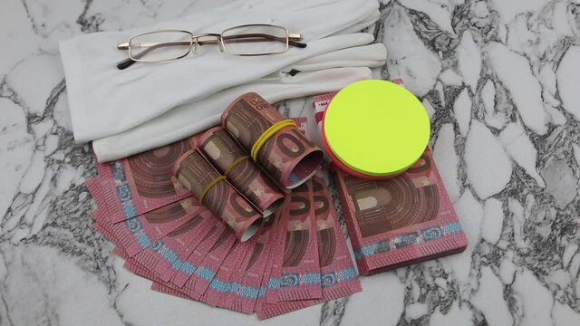 Financial or business concept with a pair of white gloves, eyeglasses and wads and fanned Euro banknotes with colorful circular memo pads over a white marble background in a flat lay view. Rotating