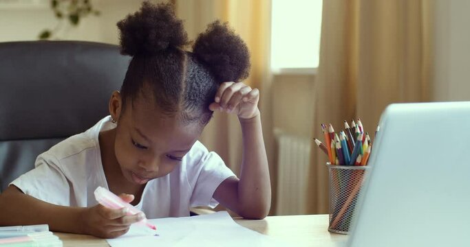 Portrait of cute African American girl child sits at home at school in kindergarten holding colorful flamasters in her hands draws with pencils on white paper makes picture remotely looking in laptop