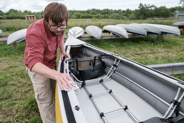 A handsome bearded man in glasses diligently wipes the kayak from water drops, on the shore, against the background of other boats.