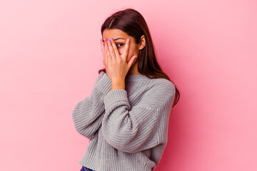 Young Indian woman isolated on pink background blink through fingers frightened and nervous.