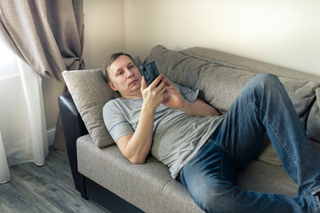 Serious man having rest using smartphone lying down on sofa at home