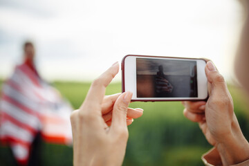 Closeup of a woman taking picture on the mobile smart phone of a man wrapped in american flag. Celebration of USA independence day at a green wheat field. Holidays concept,