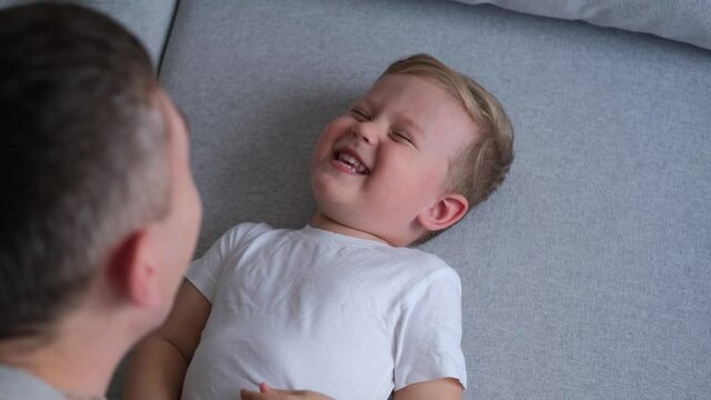 The kid lies on the couch and laughs while playing with dad. Daddy plays with his child by tickling him. Father and son are having fun.