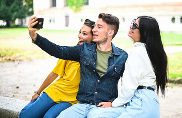 Cheerful smiling friends at the park sitting on a bench and taking selfies using a smart phone