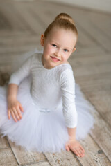 A small sweet ballerina in a white tutu and a body sitting on the birchwood floor in a light studio. Studio. Children and ballet