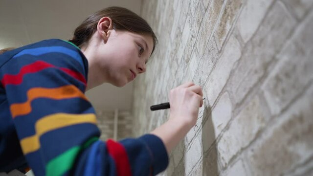 Smart girl wearing striped sweatshirt writing mathematic fractions on wallpaper with brick wall print. Low angle pupil doing homework in unusual way. Concept of education