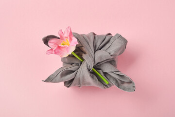 Spring eco-friendly gift wrapped in grey textile with one pink tulip flowers decor on pink. Sustainable gift. Traditional Japanese Furoshiki style. View from above.