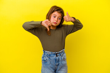Little caucasian girl isolated on yellow background showing thumbs up and thumbs down, difficult...