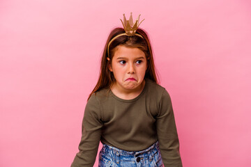 Little princess girl with crown isolated on pink background shrugs shoulders and open eyes confused.