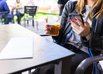 Young woman, drinking a beer on an outdoor terrace, while using her smartphone.