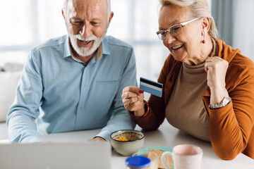 Happy mature couple shopping online over laptop while having breakfast at home