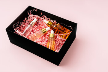 Black beauty gift box with bottles with cosmetic serum and shredded pink paper aqainst pink background. Copy space, front view.