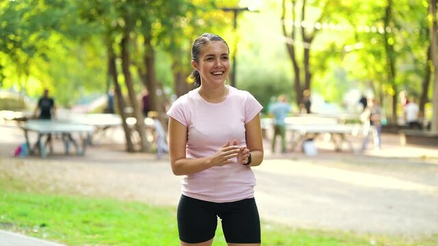 Woman with ponytail wearing pink t-shirt and black shorts laughing in summer park, blurred people on background. Active female training outside on sunny day. Concept of sport