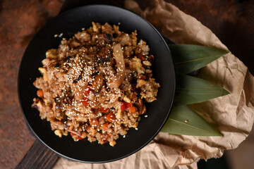 top view of hot second courses of Japanese cuisine. Chahan rice with white sesame seeds, mussels and vegetables in a black bowl on a slate board with bamboo leaves on a brown background