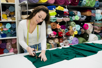 Obraz na płótnie Canvas Young brunette woman seller cutting fabrics in textile store and taking orders by smartphone. Seamstress using scissors to trim the material on work table. Drapery shop. Small Business Concept
