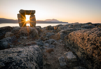 Sunset through Inuksuk in rough arctic landscape. Sunstar on the horizon in the fjord of...
