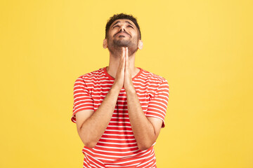 Hopeless desperate man with beard in striped t-shirt folding hands in pray looking up, communicating with god, asking for forgiveness and blessing. Indoor studio shot isolated on yellow background