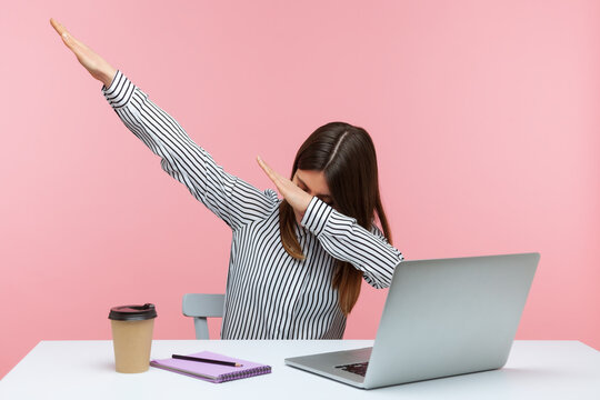 Extremely excited happy woman office worker showing dab dance gesture, performing internet meme of success, sitting at workplace with laptop. Indoor studio shot isolated on pink background