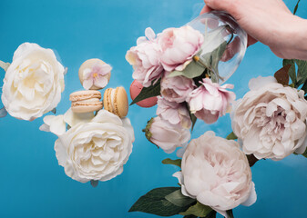 blurry hand holding glass with peons and flowers and macaroons on blue background 