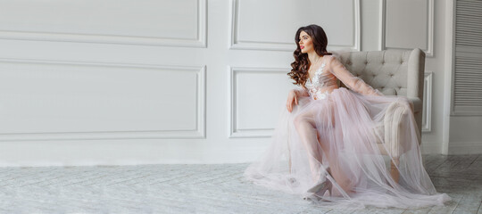 A bride in a white interior in a beautiful boudoir semi-transparent dress. She is sitting on the sofa, she has a beautiful wedding hairstyle and makeup