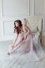 A bride in a white interior in a beautiful boudoir semi-transparent dress. She sits on the couch and puts on her shoes