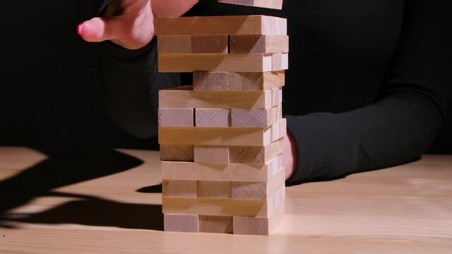 The girl skillfully plays Jenga while sitting at the table. The girl's hands carefully remove the wooden blocks and place them on the top of the tower without destroying it. Slow motion. Close up.