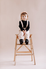 smiling little baby girl in beige jumpsuit and flower wreath on her head standing on a ladder in the studio. cute child isolated on beige background. Concept of the happy childhood.