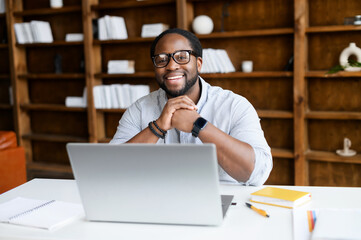 Cheerful African-American guy in smart casual wear sits at the desk with a laptop, staring into a camera with a friendly toothy smile, a biracial office employee on the workplace