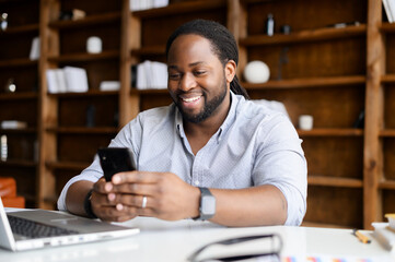 Smiling African-American man wearing smart casual shirt is using smartphone on the workplace at office, a happy mixed-race guy texting messages, enjoying chatting in social networks