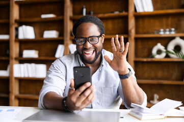 Overjoyed African-American guy using a smartphone for video call sitting at the office, a...