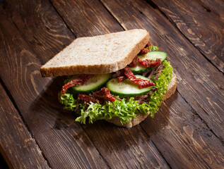 beef sandwich with green lettuce leaves, dry tomatoes and fresh cucumbers, at rustic table background