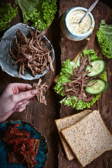 hands preparing sandwich with meat, dark bread, fresh green salad, dry tomatoes and fresh cucumbers on rustic wooden table surface