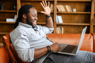 Cheerful African-American man is using a laptop for video communication with friends, family or coworkers, a guy is lying on the sofa, waving hand, greeting online participants, side view