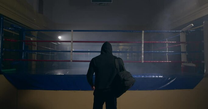 Back shot of confident athletic man entering empty gym putting sports bag down on boxing ring. Male boxer before training.