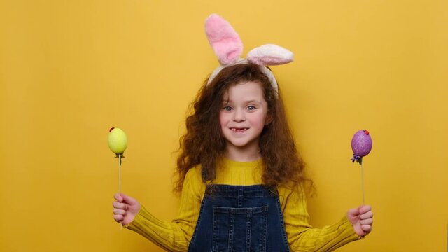 Portrait of surprised little kid girl in pink bunny fluffy ears holding two colored eggs, being symbol of religious holiday, isolated on yellow background. Childhood lifestyle and Happy Easter concept
