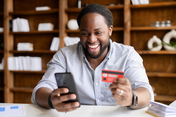 Excited African-American guy is using a smartphone and a credit card for shopping online, happy...