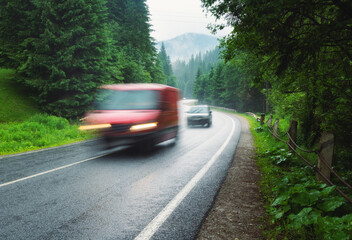 Blurred cars on the road in foggy forest in rainy day in summer. Beautiful mountain roadway, trees, green foliage in fog. Landscape with red car in motion, road through the woods. Travel. Road trip	