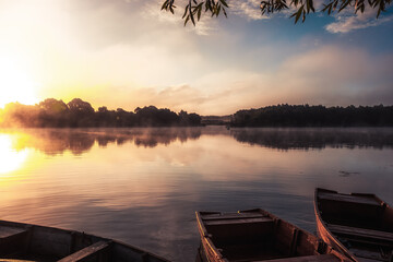 Rural countryside morning foggy scenery with boats on river with tranquil mirror water and reflections during sunrise in warm colors during golden hour