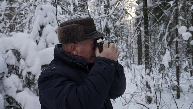old man is taking photo of snowy forest at winter day, photographing nature by camera, standing alone