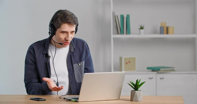 Caucasian man in headphones speaks on video call makes online conference chat sitting at table answers on questionable information says no feels shock surprise from what he heard spreads arms to sides