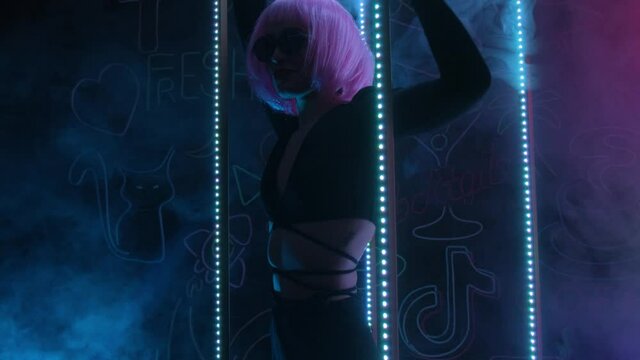Hipster girl with glasses and a pink wig in a dark neon room with smoke. The girl licks the candy.