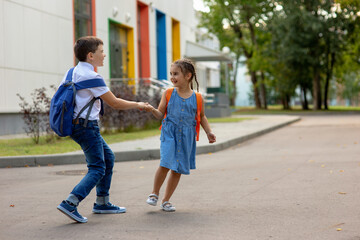 two schoolchildren, a little girl and a brother with backpacks hold hands, play and have fun near the school building